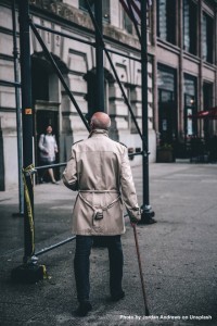 älterer Mann mit Spazierstock geht in einer Großstadt vor einem Gerüst spazieren.