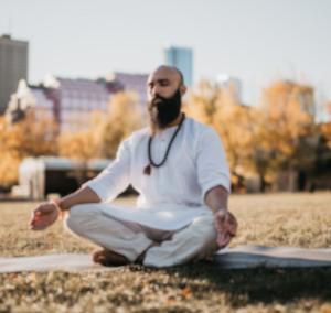 Meditation ist ein der bekanntesten Formen um Entspannung zu erreichen. Dieser Mann meditiert in einem Park vor der Skyline einer Großstadt. Im Schneidersitz auf einer Matte sitzend.