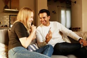 Ein junges Paar sitz auf einer Couch. Spricht glücklich miteinander. Er zeigt ihr etwas auf dem Smartphone. Die Kraft der Ehrlichkeit-Beziehungstipps