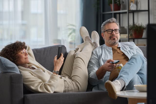 Paar 2025 sitzt nebeneinander auf dem Sofa, einer am Handy , der andere mit der Fernbedienung vom TV– Symbol für den neuen Paar-Trend: Nähe muss neu definiert werden.