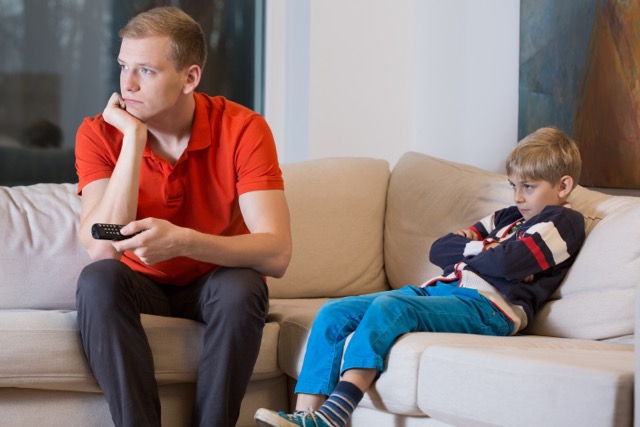 Loyalitätskonflikt bei Kindern: Junge sitzt abends wach auf dem Sofa neben seinem Vater und langweilt sich. Vater ist in Gedanken und anderweitig beschäftigt.